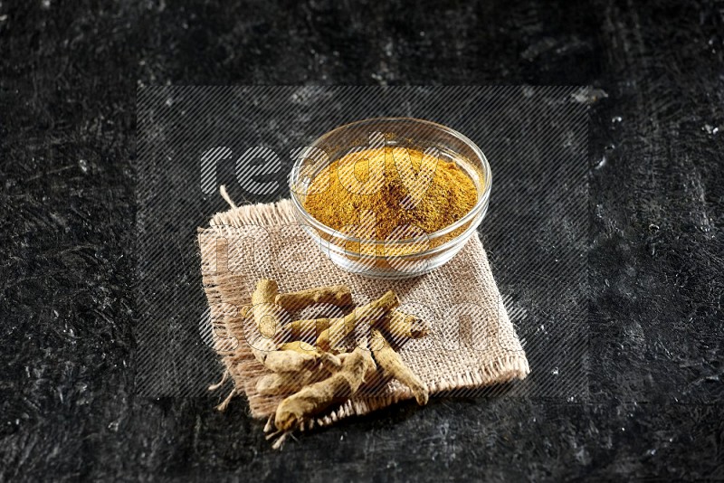 A glass bowl full of turmeric powder with dried turmeric fingers on a burlap fabric on textured black flooring