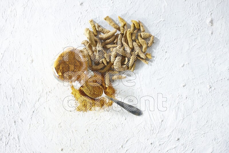 A flipped glass spice jar and metal spoon full of turmeric powder and powder spilled out of it with dried whole fingers on textured white flooring