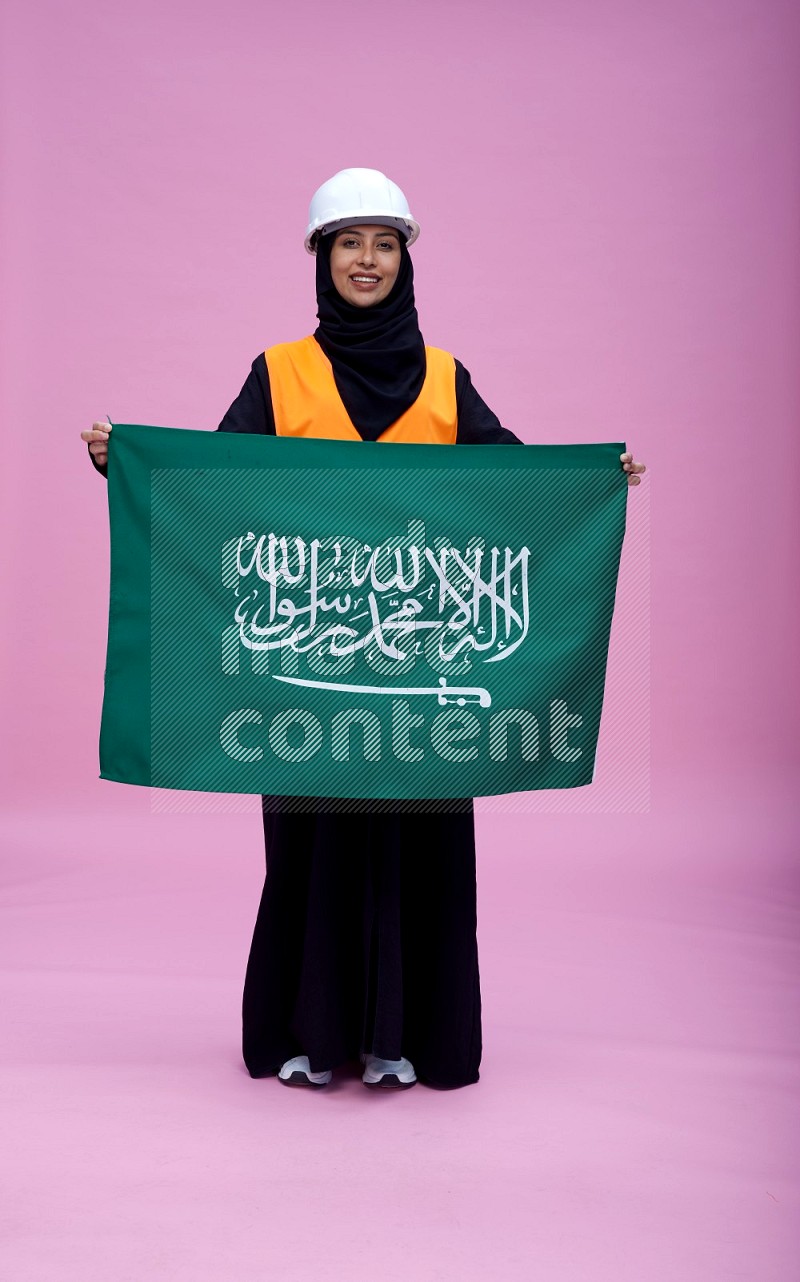 Saudi woman wearing Abaya with engineer vest and helmet standing holding Saudi flag on pink background