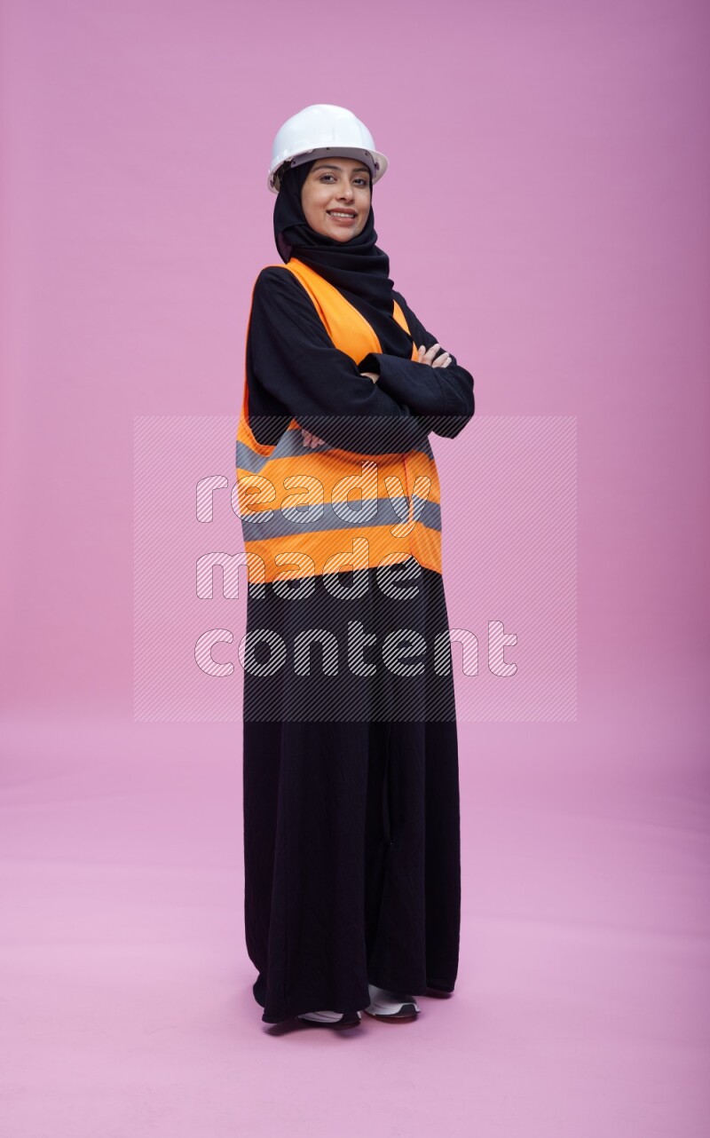 Saudi woman wearing Abaya with engineer vest and helmet standing with crossed arms on pink background