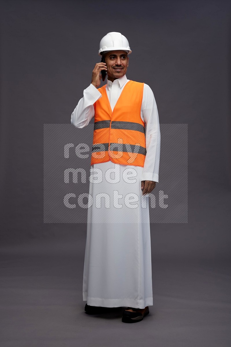 Saudi man wearing thob with engineer vest standing talking on phone on gray background