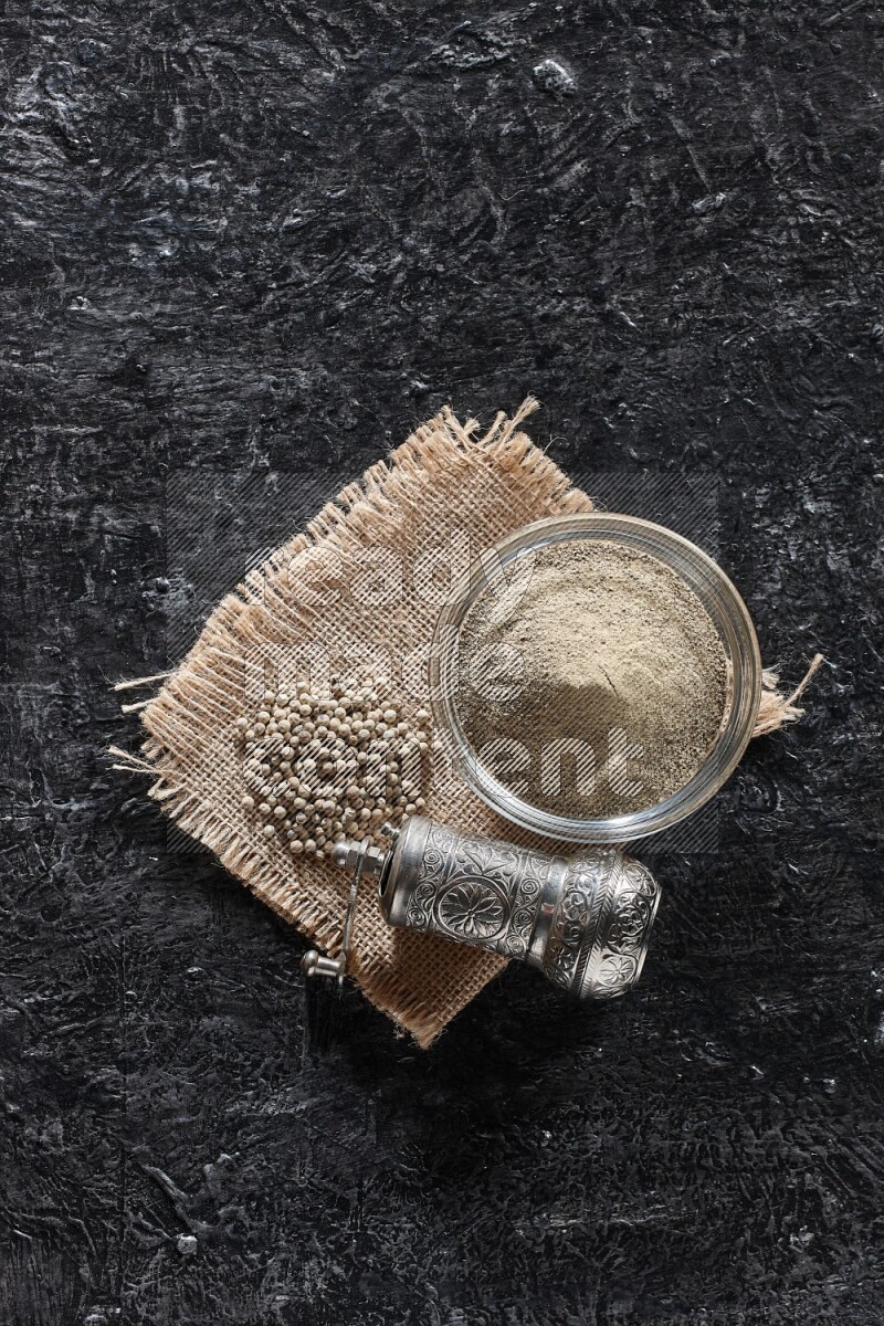 A glass bowl full of white pepper powder with white pepper beads on a burlap piece of fabric and a metal grinder on textured black flooring