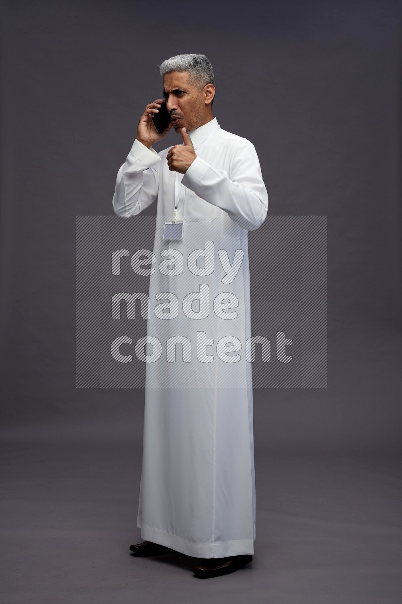 Saudi man wearing thob with neck strap employee badge standing talking on phone on gray background