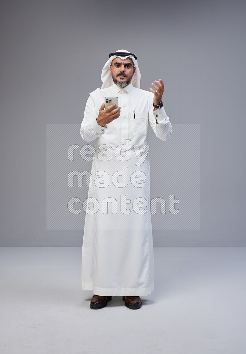 Saudi man Wearing Thob and white Shomag standing texting on phone on Gray background