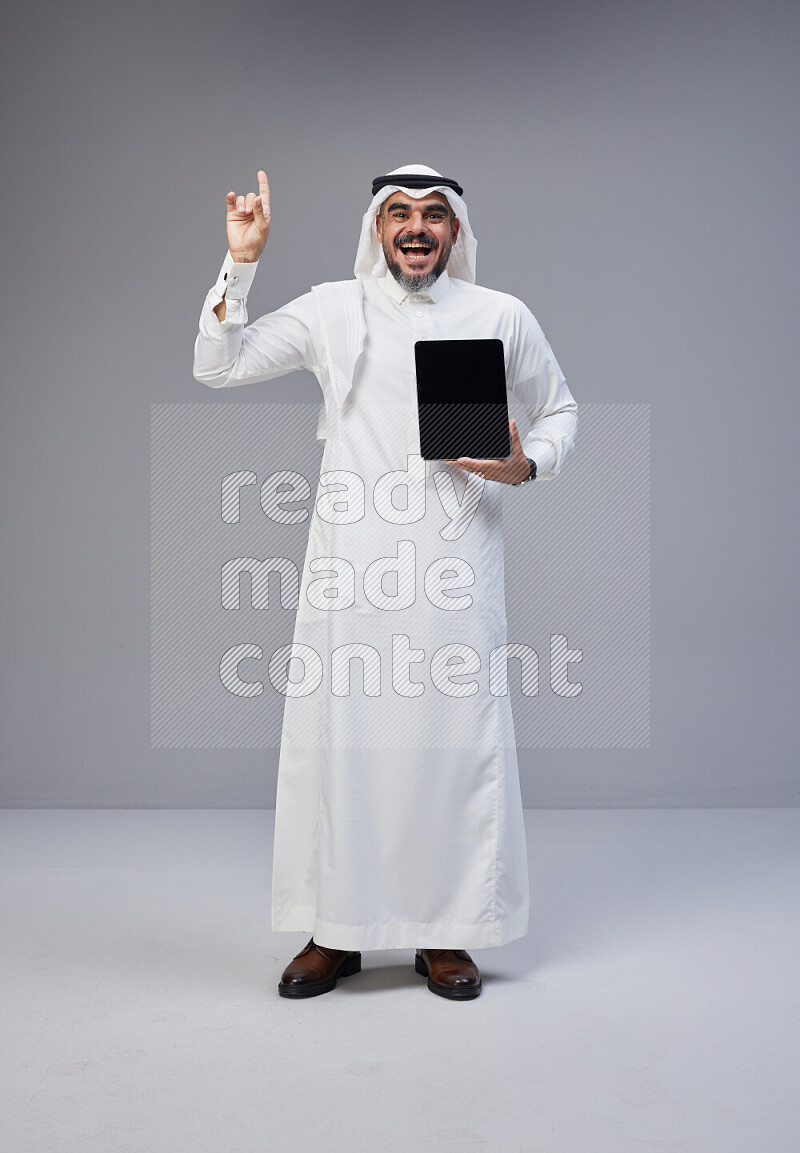 Saudi man Wearing Thob and white Shomag standing showing tablet to camera on Gray background