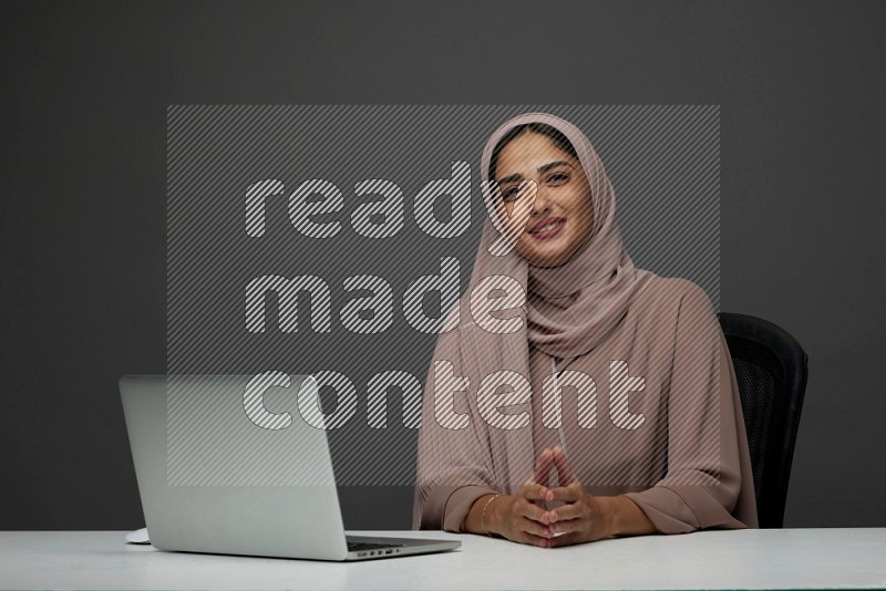 A Saudi woman Setting on her desk on a Gray Background wearing Brown Abaya with Hijab