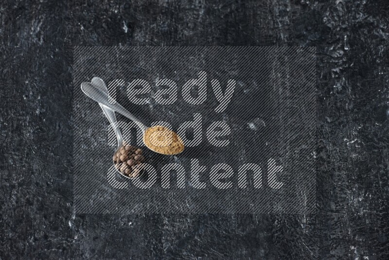 ملعقتان معدنيتان ممتلئتان ببودرة بهار الفلفل الحلو وحبوبه على خلفية سوداء