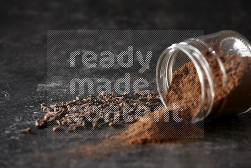 A flipped glass jar full of cloves powder with cloves spread on a textured black flooring