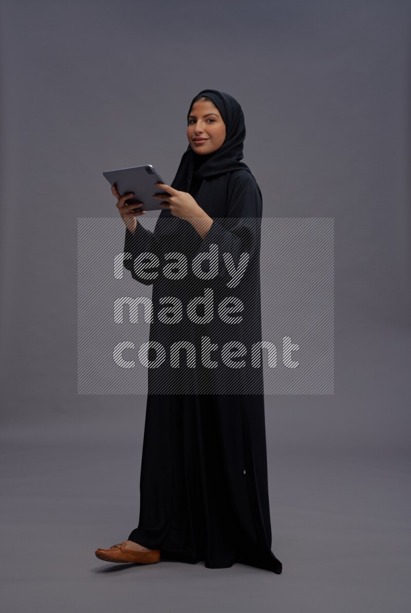 Saudi woman wearing Abaya standing working on tablet on gray background