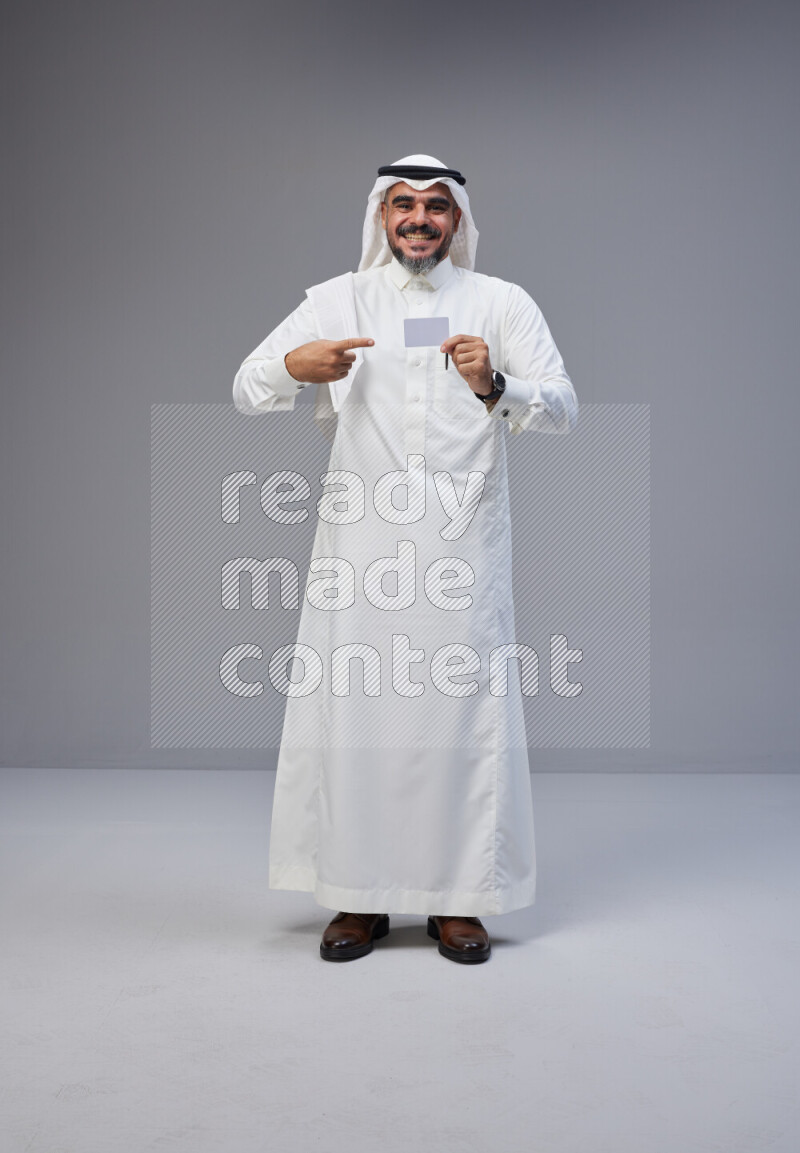 Saudi man Wearing Thob and white Shomag standing holding ATM card on Gray background