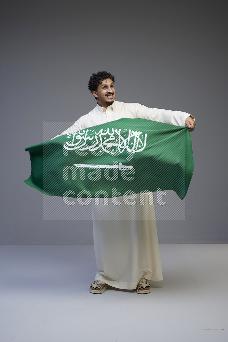 A saudi man standing wearing thob holding big saudi flag on gray background