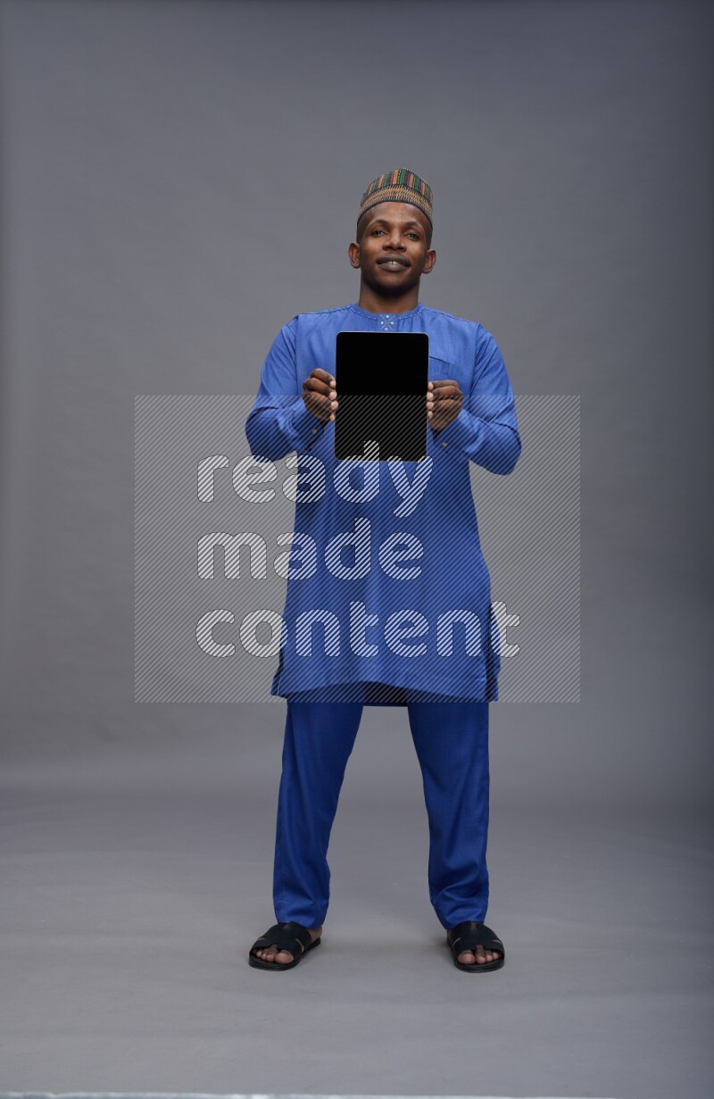 Man wearing Nigerian outfit standing showing tablet to camera on gray background