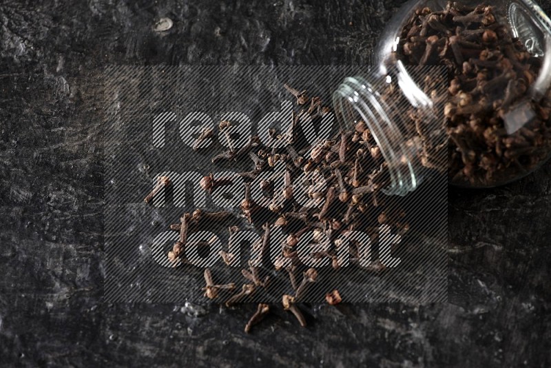 A flipped glass spice jar full of cloves and cloves came out of it on textured black flooring