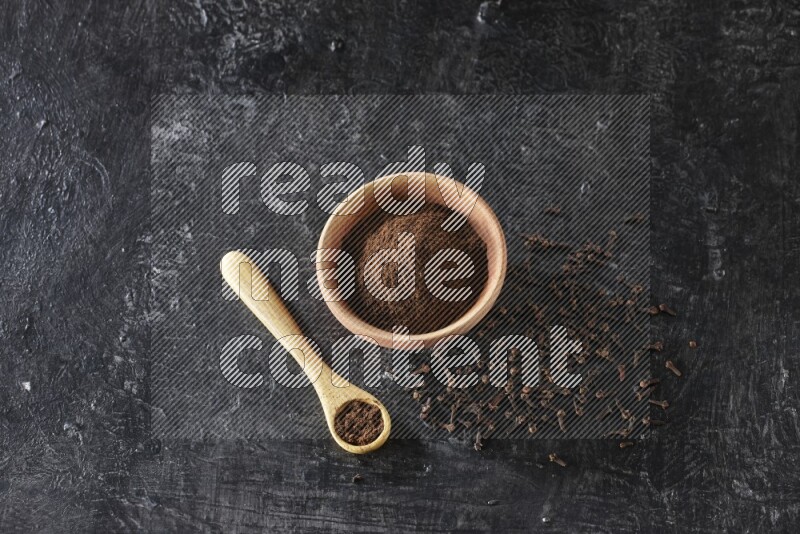 A wooden bowl and wooden spoon full of cloves powder with spreaded cloves on textured black flooring