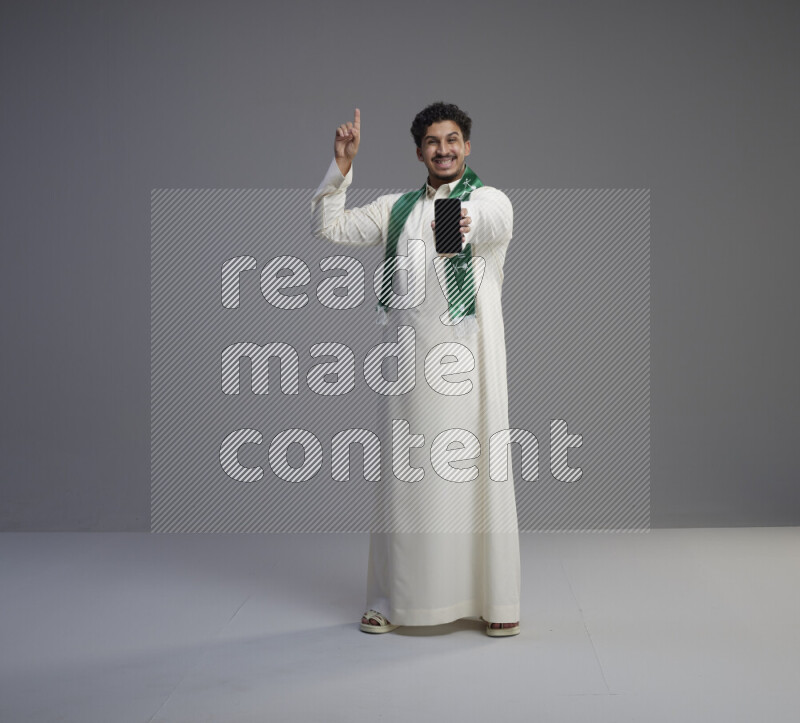 A Saudi man standing wearing thob and Saudi flag scarf showing phone to camera on gray background