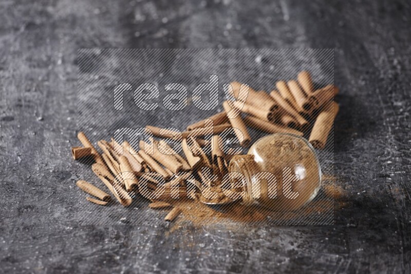 Herbal glass jar full cinnamon powder flipped and a metal spoon full of powder surrounded by cinnamon sticks on textured black background in different angles