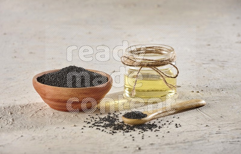 وعاء خشبية وملعقة مليئان ببذور حبة البركة مع وعاء زجاجي من زيت حبة البركة على أرضية بيضاء