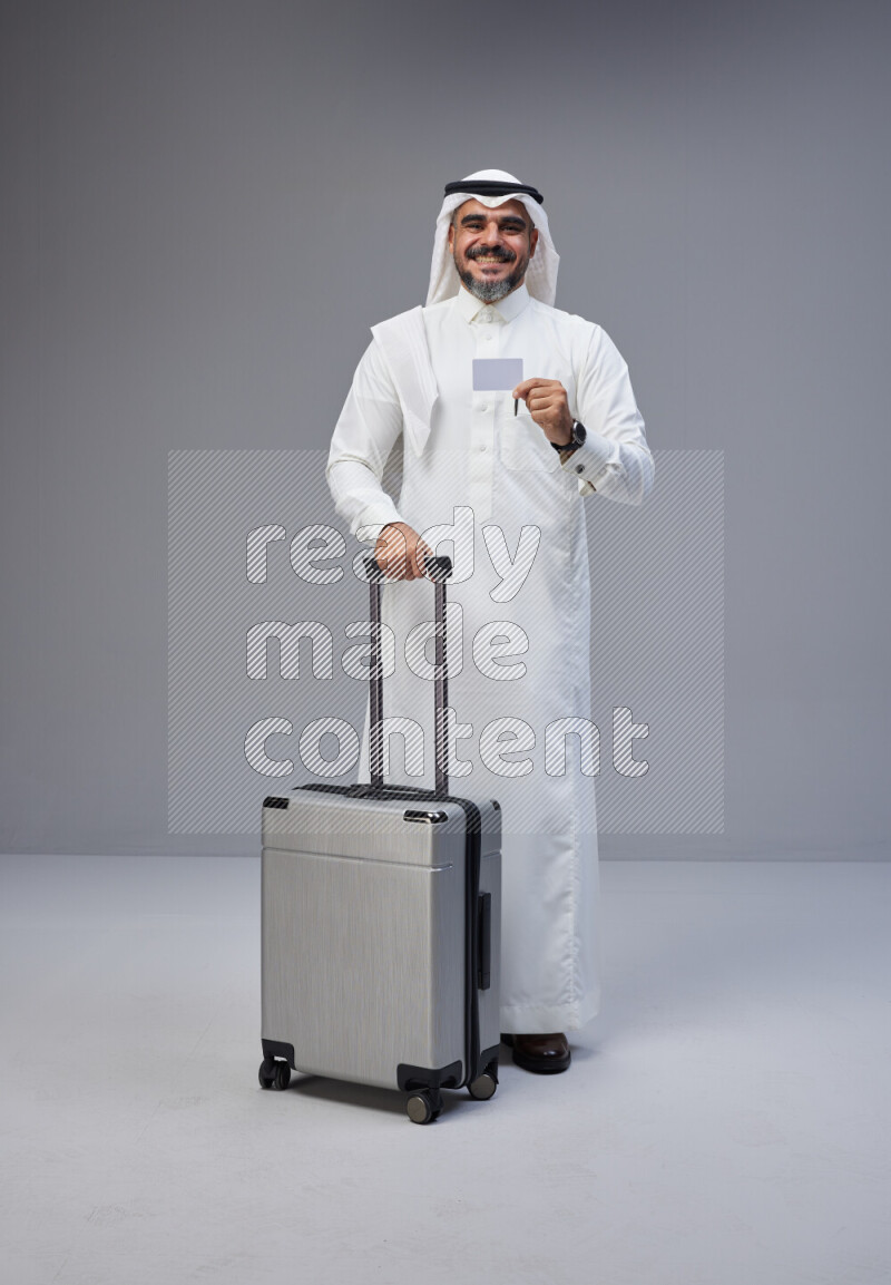 Saudi man wearing Thob and white Shomag standing holding Travel bag and ATM card on Gray background