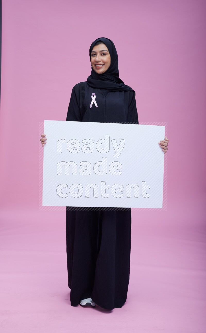 Saudi woman wearing pink ribbon on Abaya standing holding board on pink background