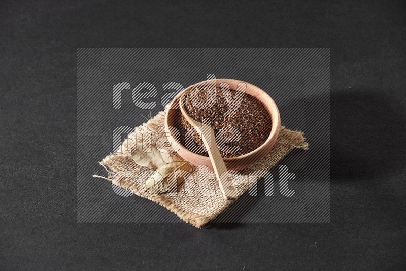A wooden bowl full of flaxseeds with wooden spoon full of the seeds on it on burlap fabric on a black flooring