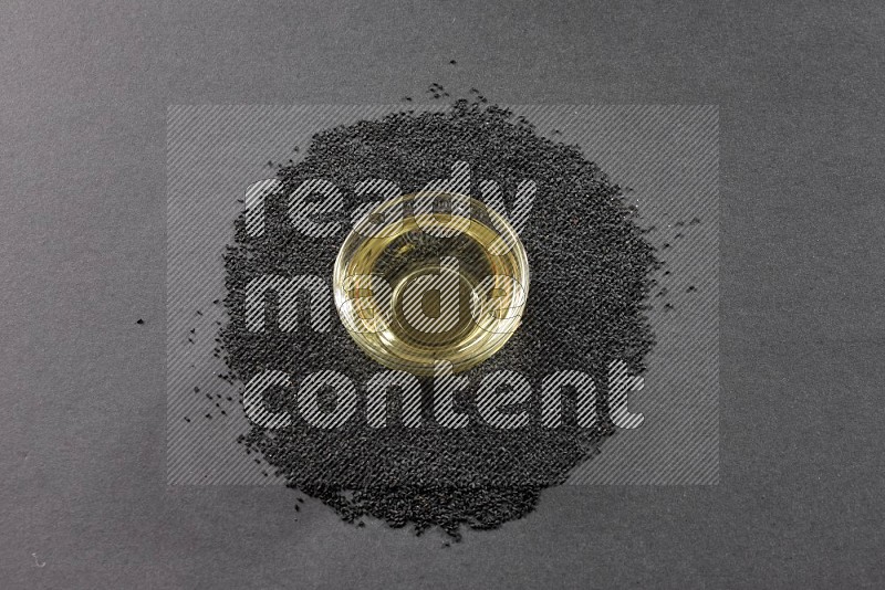 A glass bowl full of black seeds oil surrounded by the seeds on a black flooring