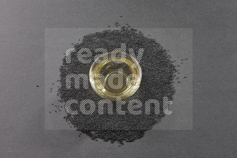 A glass bowl full of black seeds oil surrounded by the seeds on a black flooring