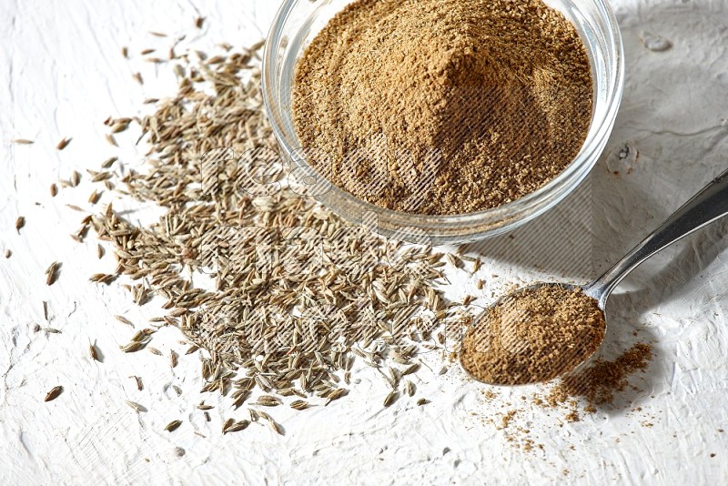 A glass bowl and metal spoon full of cumin powder and cumin seeds underneath it on textured white flooring