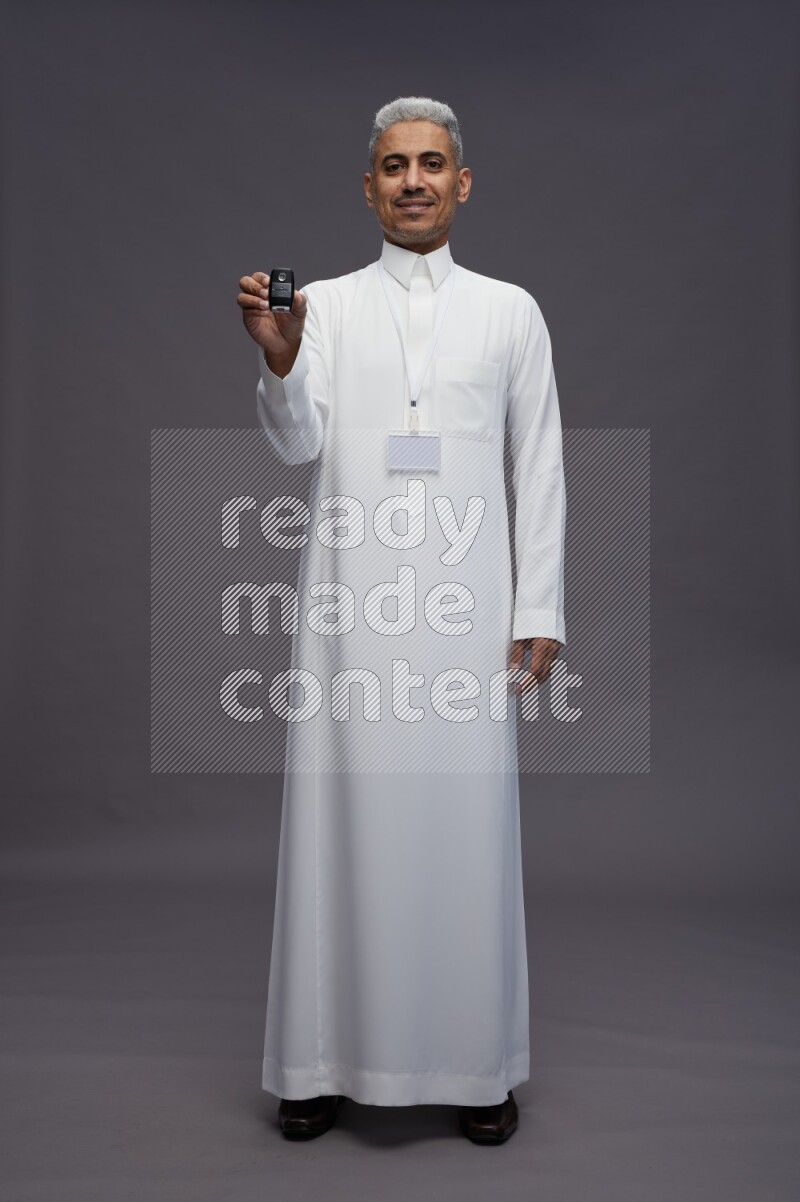 Saudi man wearing thob with neck strap employee badge standing holding key car on gray background