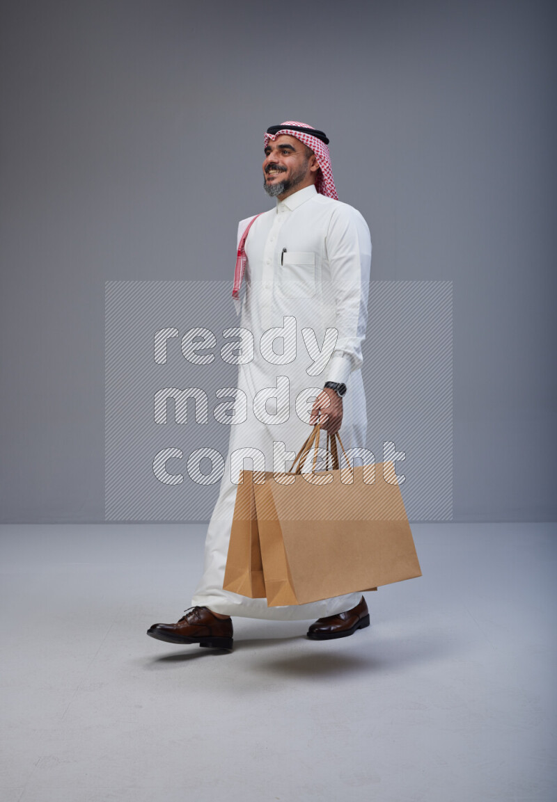 Saudi man Wearing Thob and red Shomag standing holding shopping bag on Gray background