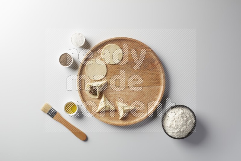 two closed sambosas and one open sambosa filled with meat while flour, salt, black pepper and oil with oil brush aside in a wooden dish on a white background