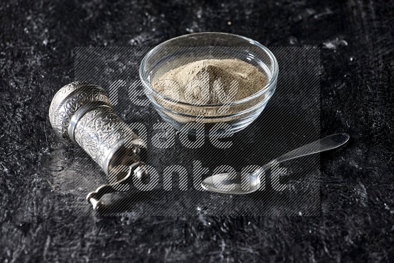 وعاء زجاجي ممتلئ ببودرة الفلفل الأبيض مع حبوب فلفل، مطحنة معدنية وملعقة معدنية على أرضية سوداء