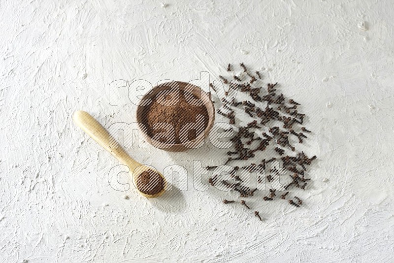 A wooden bowl and wooden spoon full of cloves powder with cloves spread on textured white flooring