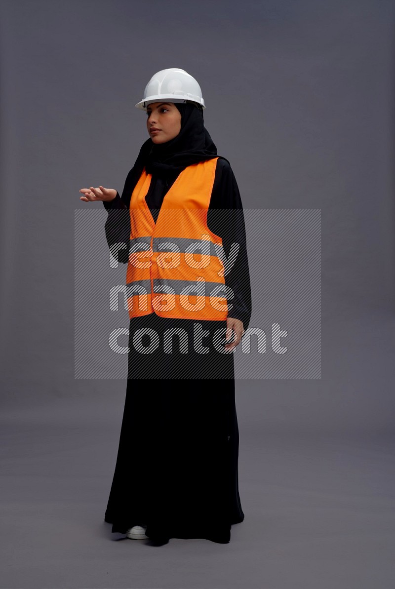 Saudi woman wearing Abaya with engineer vest standing interacting with the camera on gray background