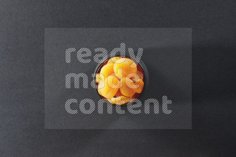 A black pottery bowl full of dried apricots on a black background in different angles