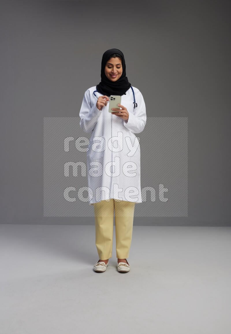 Saudi woman wearing lab coat with stethoscope standing texting on phone on Gray background