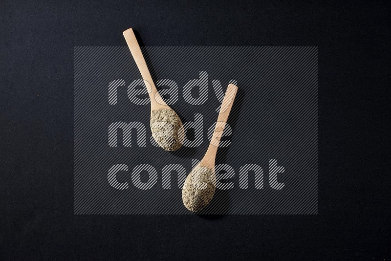 2 Wooden spoons full of cardamom powder on black flooring