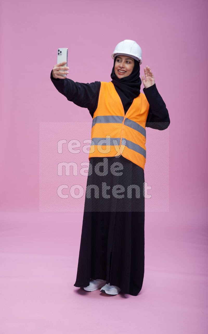 Saudi woman wearing Abaya with engineer vest and helmet standing taking selfie on pink background