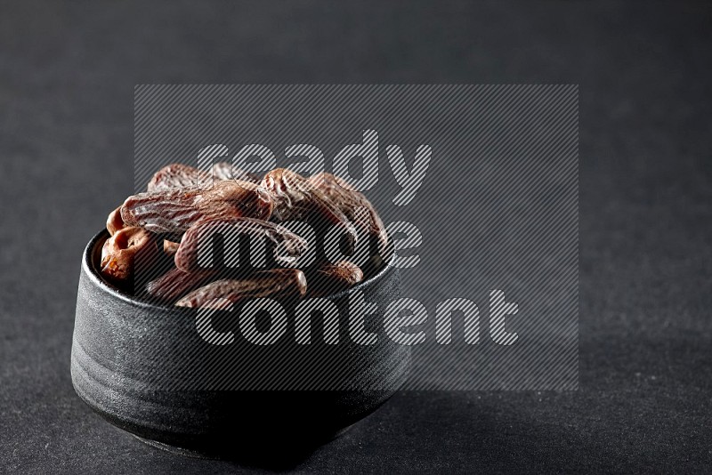 A black pottery bowl full of dried dates on a black background in different angles
