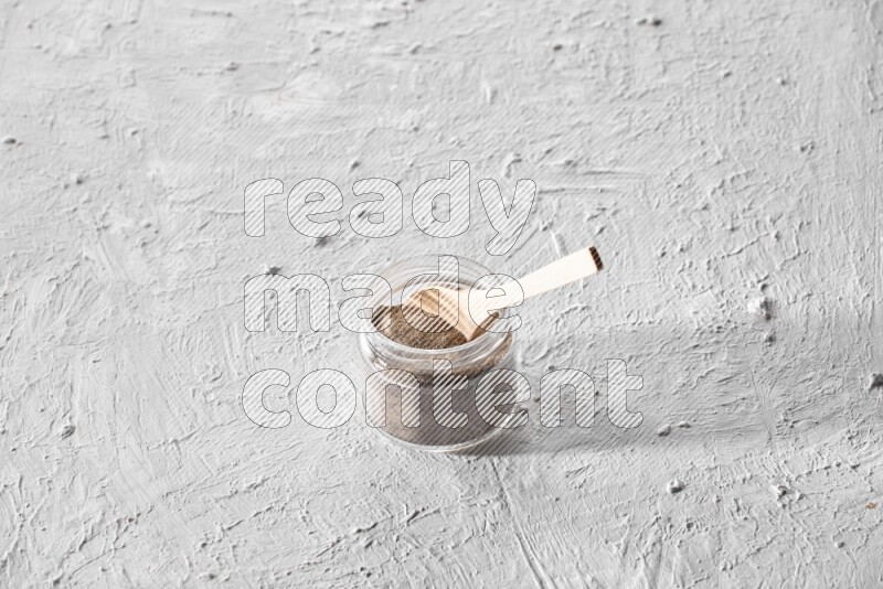 A glass jar full of black pepper powder with a wooden spoon on a textured white flooring