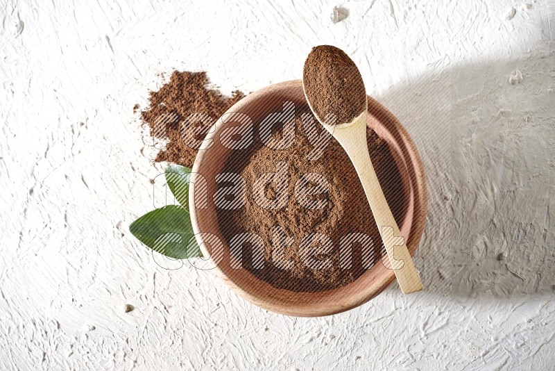 A wooden bowl and a wooden spoon full of cloves powder on a textured white flooring