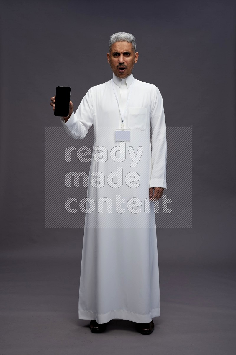 Saudi man wearing thob with neck strap employee badge standing showing phone to camera on gray background
