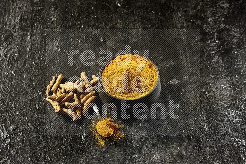 A pottery black bowl and a metal spoon full of turmeric powder with dried turmeric fingers on a textured black flooring
