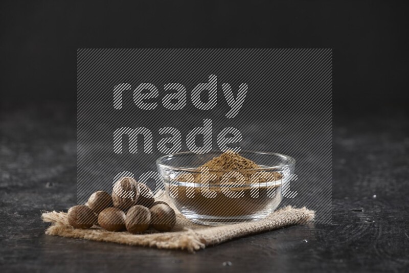 A glass bowl full of nutmeg powder with whole seeds beside it on burlap fabric on a textured black flooring