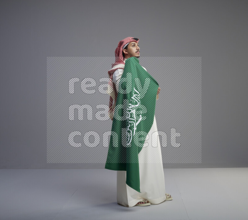 رجل سعودي يرتدي ثوب ابيض وشماغ احمر ويحمل علم كبير للسعودية