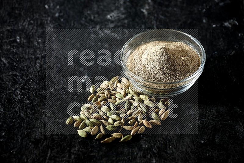 A glass bowl full of cardamom powder and cardamom seeds beside it on textured black flooring