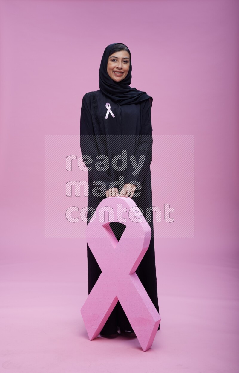 Saudi woman wearing pink ribbon on abaya standing holding awareness ribbon on plain pink background