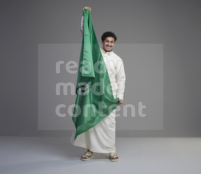 A Saudi man standing wearing thob holding big Saudi flag on gray background