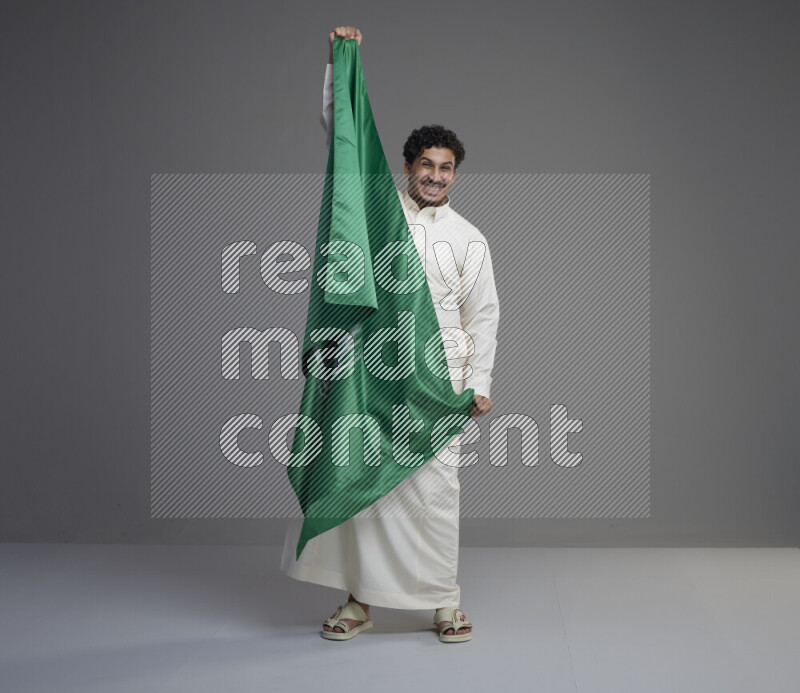 A Saudi man standing wearing thob holding big Saudi flag on gray background