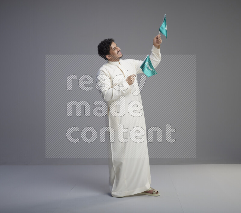 رجل سعودي يرتدي ثوب ابيض يحمل علم السعودية على خلفية رمادي