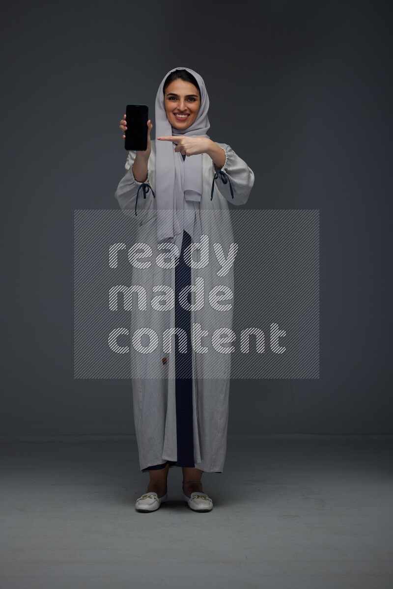 A Saudi woman wearing a light gray Abaya and head scarf standing and showing the phone's screen while holding a blue luggage eye level on a grey background